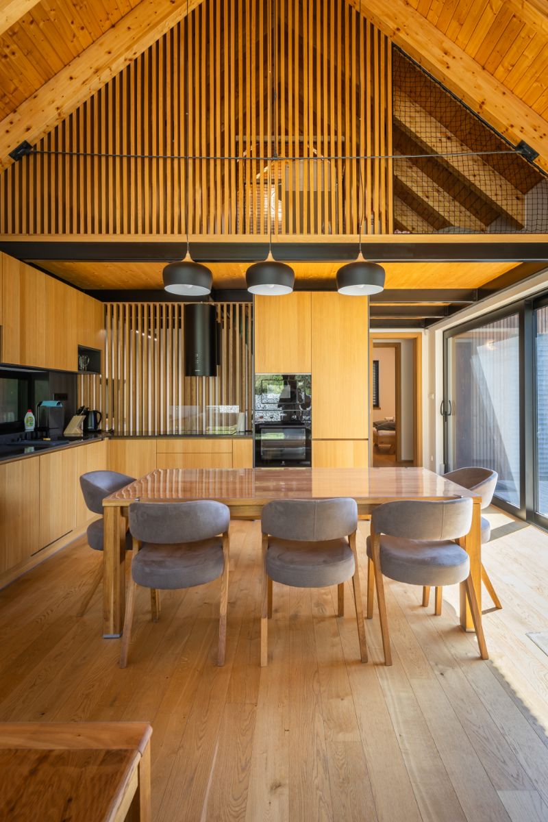 Moderná kuchyňa a jedáleň v chate s drevenými obkladmi, trámami a šedými stoličkami pri lesklom drevenom stole.