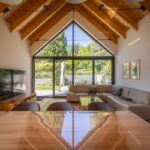 Priestranná obývacia izba s vysokým dreveným stropom, béžovou sedačkou a krbom s presklenou stenou s výhľadom do zelenej záhrady.