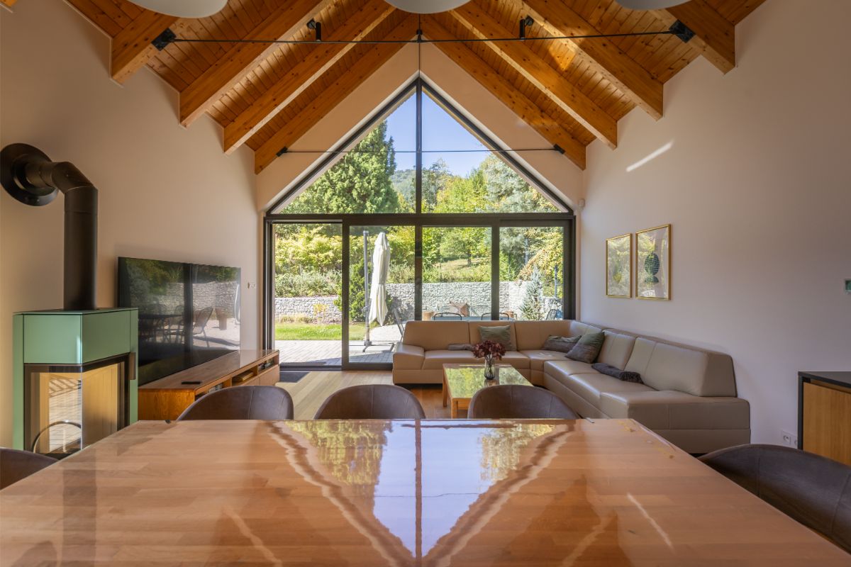 Priestranná obývacia izba s vysokým dreveným stropom, béžovou sedačkou a krbom s presklenou stenou s výhľadom do zelenej záhrady.