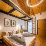 Spálňa s drevenou manželskou posteľou, drevenou podlahou a stropom, s kruhovým neónovým svetlom a rebríkom na sieťovanú galériu.