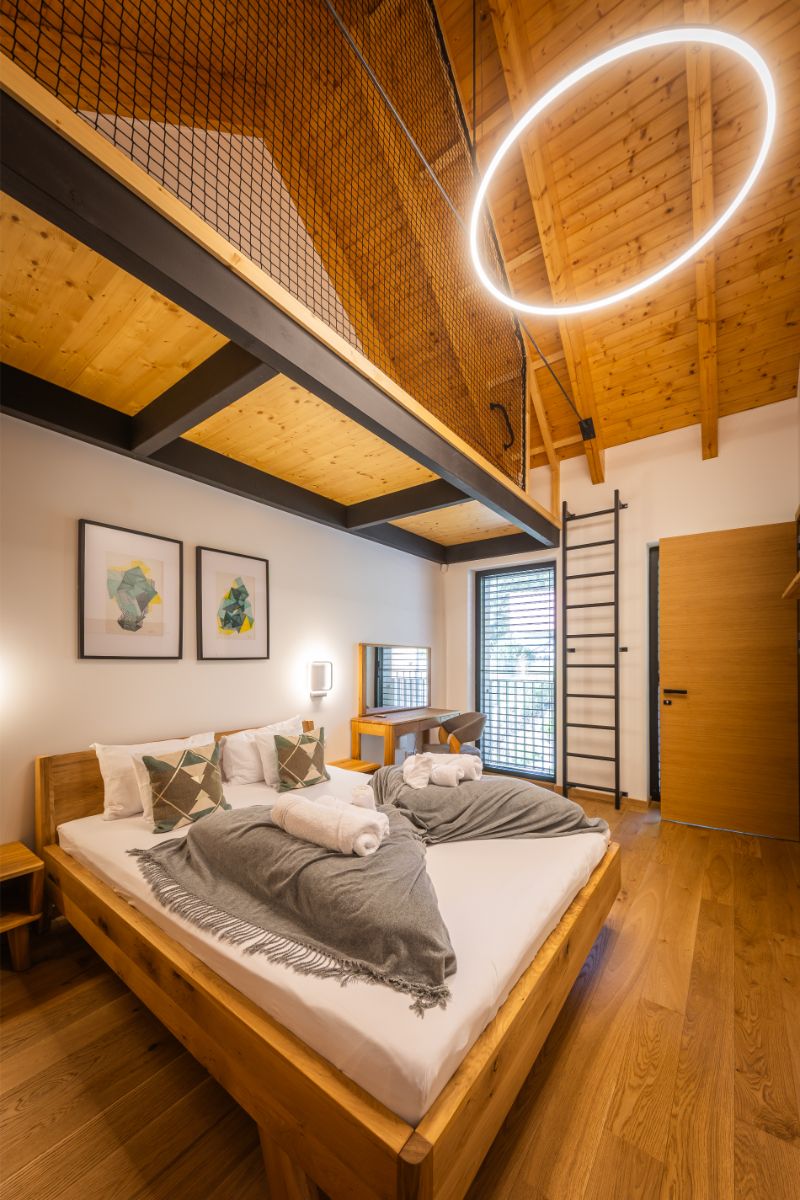Spálňa s drevenou manželskou posteľou, drevenou podlahou a stropom, s kruhovým neónovým svetlom a rebríkom na sieťovanú galériu.