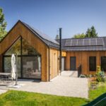 Drevená rekreačná chata s fotovoltickými panelmi na čiernej streche a veľkou terasou s dlažbou a vonkajším posedením na slnku.