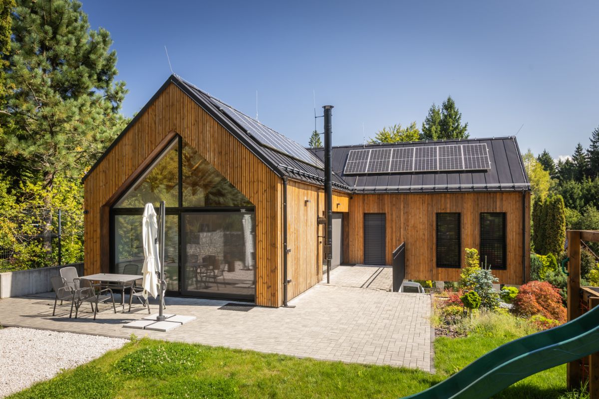 Drevená rekreačná chata s fotovoltickými panelmi na čiernej streche a veľkou terasou s dlažbou a vonkajším posedením na slnku.