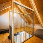 Podkrovná galéria v chate s drevenou konštrukciou a podlahou, so sieťovanou bezpečnostnou ohrádkou a drevenými trámami.