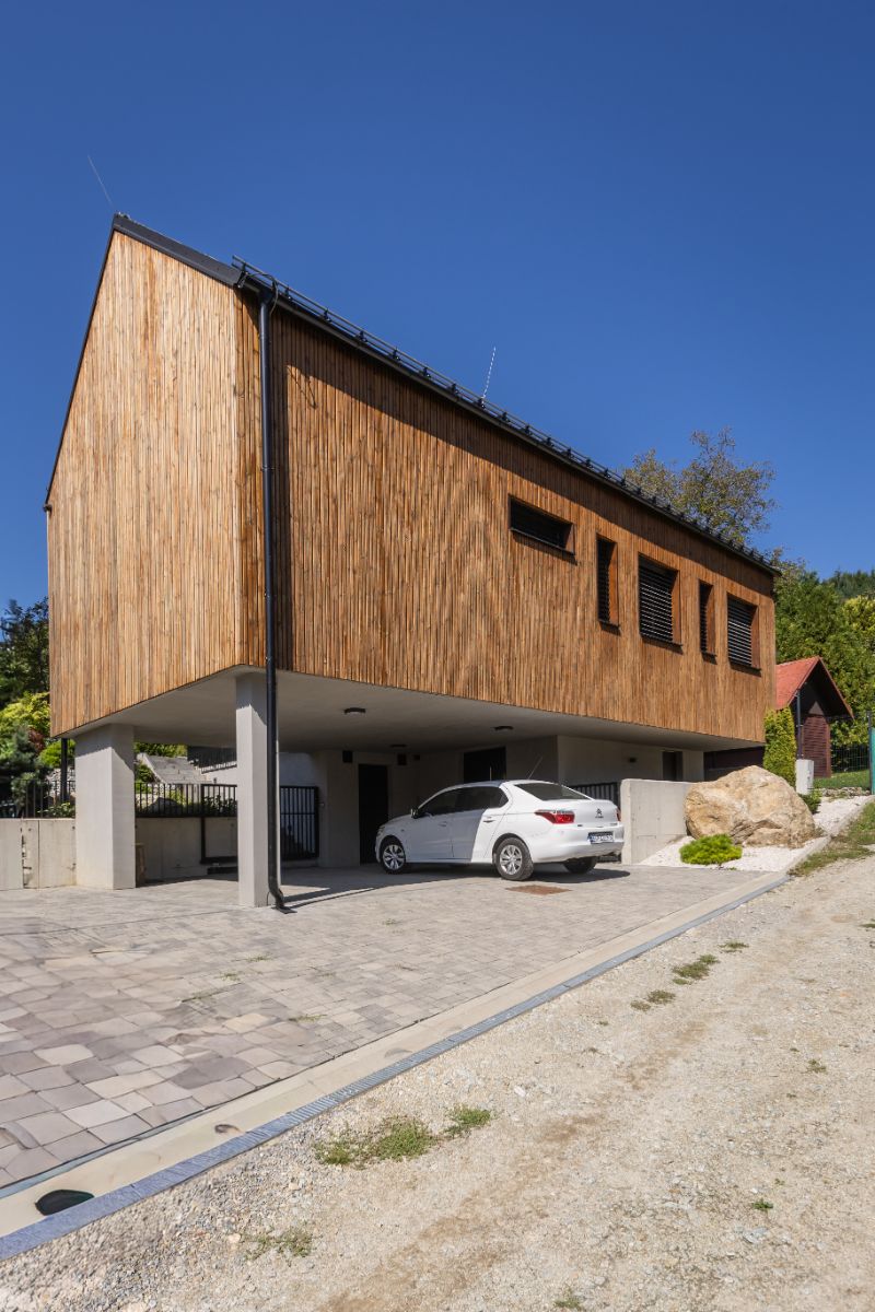 Jedno rameno horného podlažia stavby stojí na pilieroch. Vznikol tak krytý priestor na parkovanie áut. Táto časť tvorí nočné krídlo domu. Za plotom sa začína svahovitá záhrada.