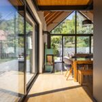 Svetlý interiér s drevenou podlahou, jedálenským kútom a moderným krbom, s veľkou presklenou stenou s výhľadom do záhrady.