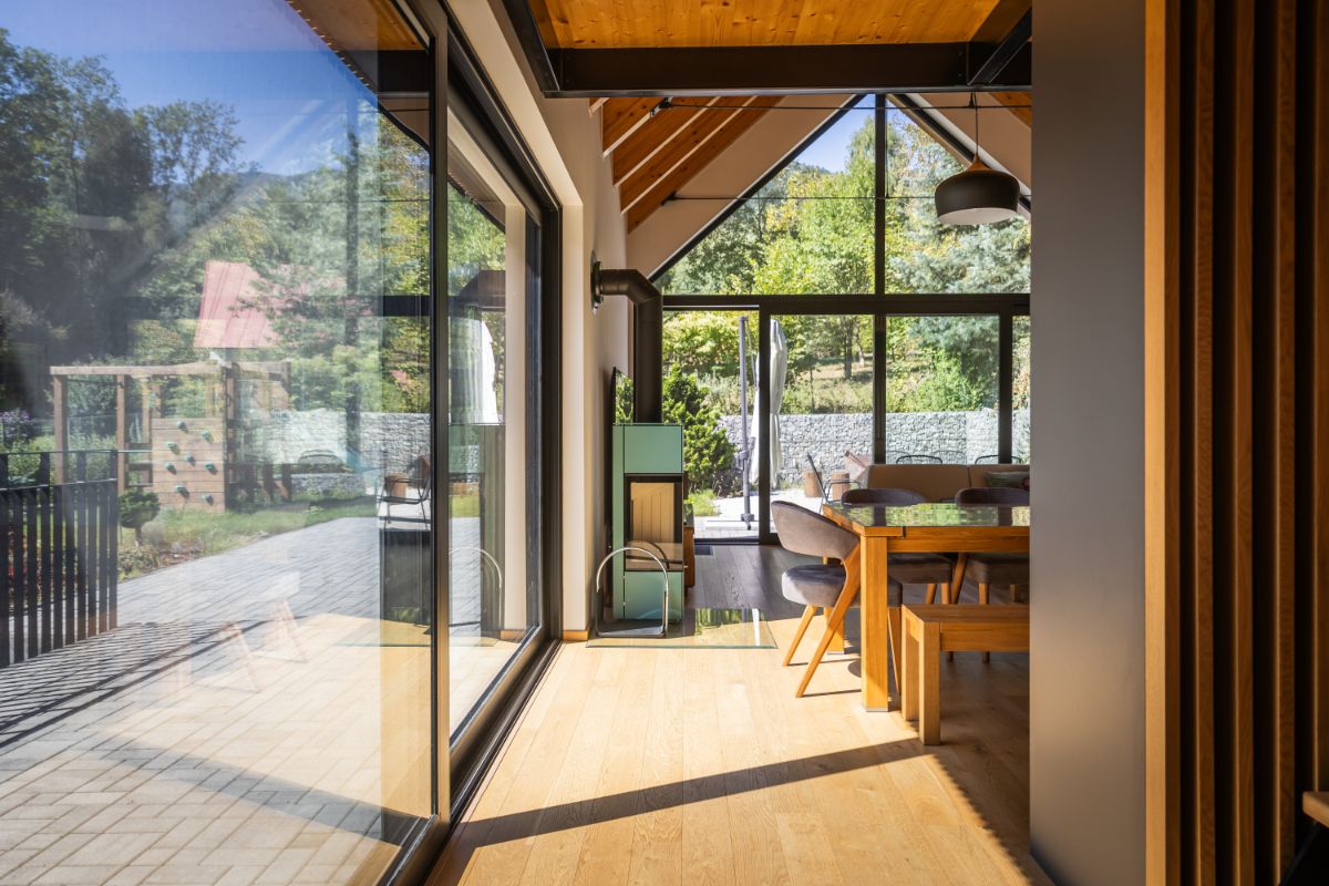 Svetlý interiér s drevenou podlahou, jedálenským kútom a moderným krbom, s veľkou presklenou stenou s výhľadom do záhrady.