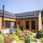 Exteriér modernej drevenej chaty s čiernou strechou, fotovoltickými panelmi a upraveným trvalkovým záhonom.