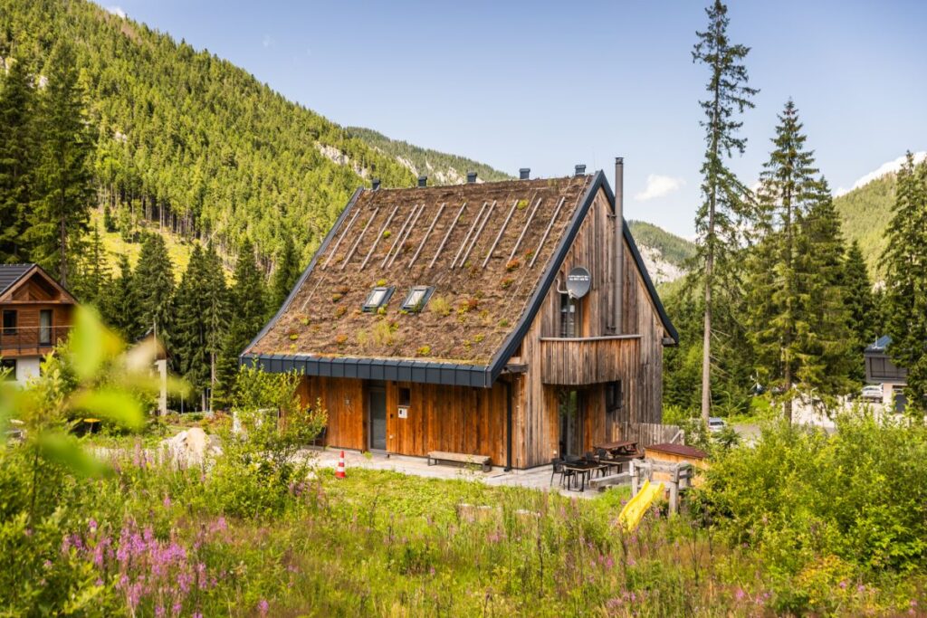 Drevená chata so šikmou zelenou strechou a priestrannou terasou v slnečnej horskej prírode.