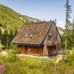 Drevená chata so šikmou zelenou strechou a priestrannou terasou v slnečnej horskej prírode.