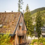 Chata s tmavým dreveným obkladom, zelenou strechou a malým balkónom s vonkajším posedením.