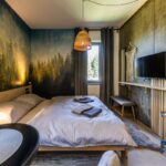 Spálňa s veľkým lôžkom, fototapetou lesa a stenami z pohľadového betónu s teplým osvetlením.