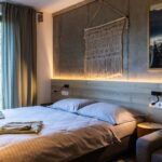 Spálňa s betónovou stenou, drevenou zástenou za posteľou a tkanou makramé dekoráciou