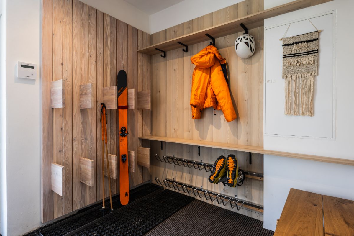 Chodba s dreveným obkladom, lyžiarskym vybavením a oranžovou bundou na vešiakoch.
