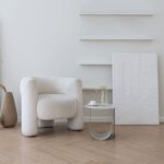 Interiér s bielym bouclé kreslom, stolíkom, abstraktným obrazom a prázdnymi policami na parketovej podlahe.