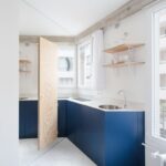 Rohová kuchynská linka s tmavomodrými skrinkami, antikorovým drezom a drevenými poličkami v minimalistickom štýle.