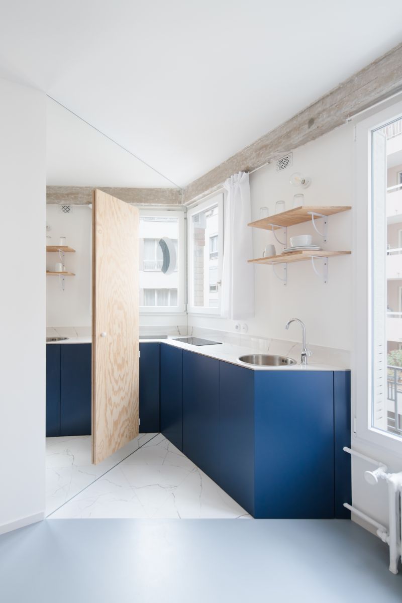 Rohová kuchynská linka s tmavomodrými skrinkami, antikorovým drezom a drevenými poličkami v minimalistickom štýle.