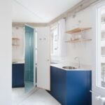 Rohová kuchynská linka s tmavomodrými skrinkami, drevenými policami a otvorenými dverami na zrkadlovej stene oproti.