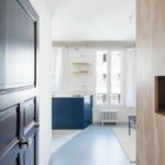 Vstupný priestor s tmavými dverami a dreveným obkladom, otvárajúci sa do svetlého priestoru s modrou podlahou a kuchyňou.