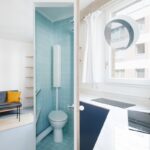 Minimalistický byt s drevenými dverami, svetlým kuchynským kútom a toaletou s tyrkysovými štvorcovými dlaždicami.