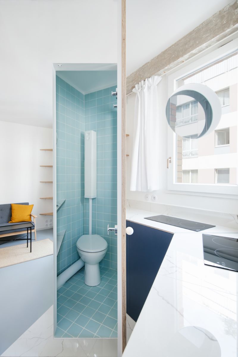 Minimalistický byt s drevenými dverami, svetlým kuchynským kútom a toaletou s tyrkysovými štvorcovými dlaždicami.