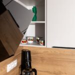 Otvorená biela policová skrinka nad drevenou zástenou s dekoratívnym riadom a čiernym kávovarom na linke.