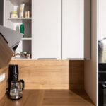 Horné biele kuchynské skrinky s čiernymi úchytkami umiestnené nad dreveným obkladom steny v čistých líniách.