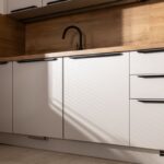 Biele kuchynské skrinky s čiernym drezom a batériou a drevenou zástenou.