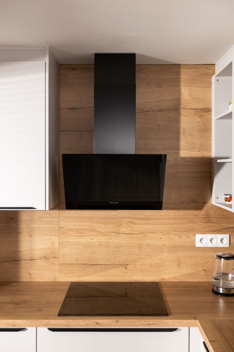 Čierny moderný digestor nad indukčnou varnou doskou s dreveným stenovým panelom v pozadí.