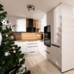 Rohová biela kuchynská zostava s vysokou skriňou na spotrebiče, dreveným obkladom a svetlou dlažbou na podlahe.