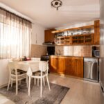 Tradičná kuchyňa s drevenou linkou, bielym jedálenským kútom a oknom s priesvitnou záclonou.