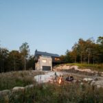 Moderná dvojpodlažná chata s tmavým a svetlým dreveným obkladom, s posedením pri ohni na skalách.