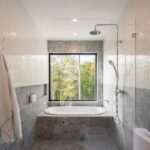 Moderná kúpeľňa s vaňou s výhľadom do prírody, obložená bielymi kachličkami a sivou žulou.