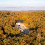 Letecký pohľad na modernú čiernobielu chatu v jesennom listnatom lese s okolitými kopcami.