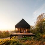 Malá moderná čierna chata s presklenou stenou a terasou s lavičkami uprostred jesennej prírody.