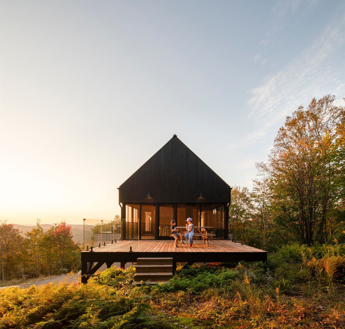 Malá moderná čierna chata s presklenou stenou a terasou s lavičkami uprostred jesennej prírody.