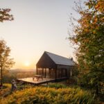 Malá moderná čierna chata s veľkou drevenou terasou a skleneným zábradlím pri západe slnka v lese.