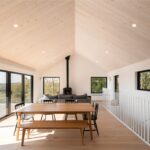 Interiér obývačky s vysokým šikmým stropom s bledým dreveným obkladom, krbom a veľkými oknami.