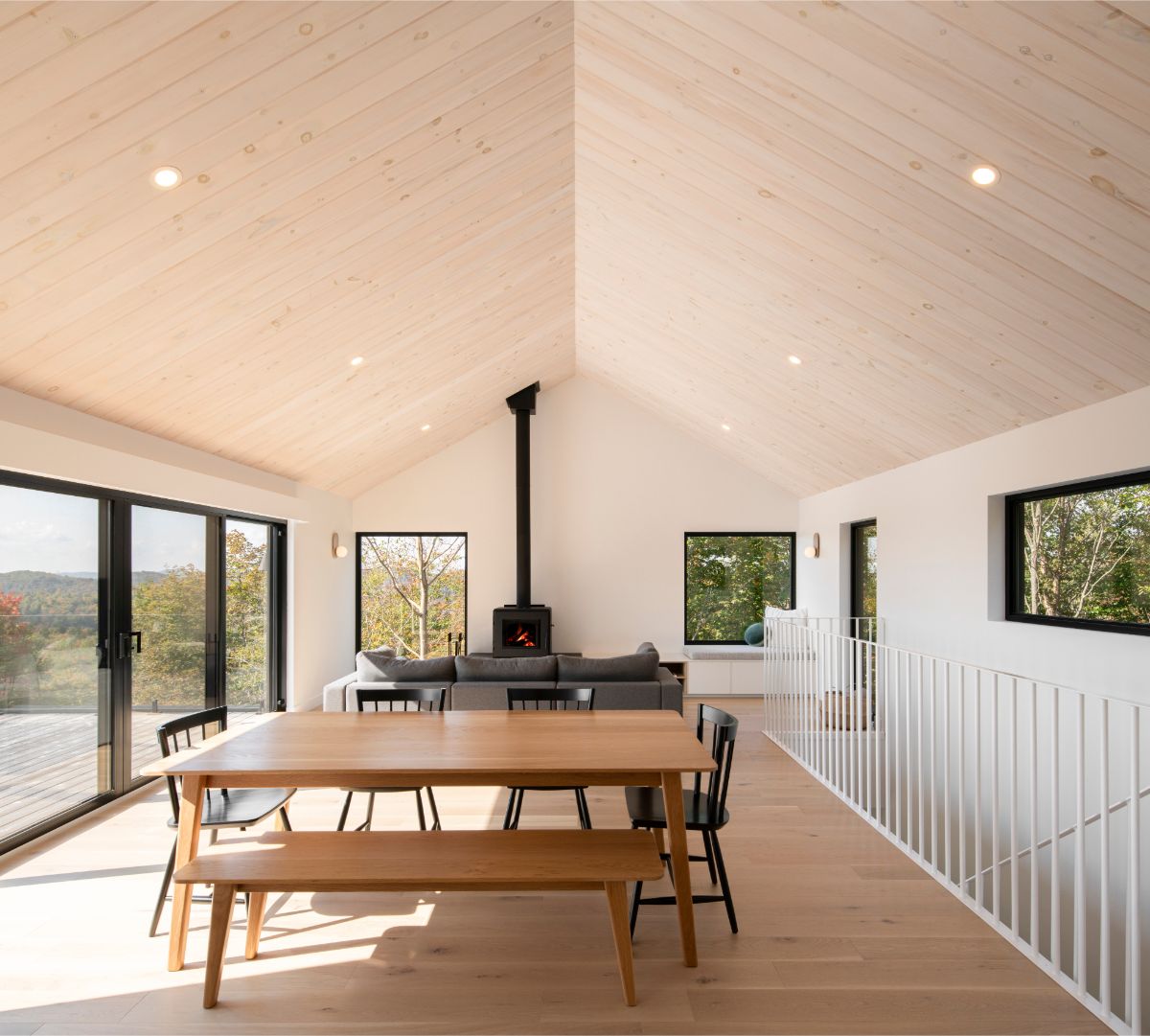 Interiér obývačky s vysokým šikmým stropom s bledým dreveným obkladom, krbom a veľkými oknami.