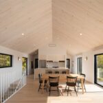 Otvorený priestor s bledým dreveným stropom, jedálenským stolom, kuchyňou a bielym zábradlím schodiska.