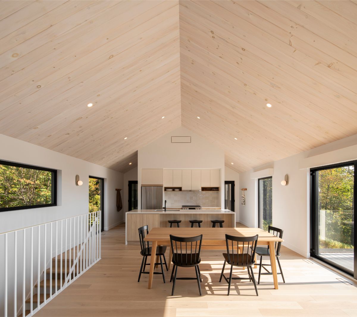 Otvorený priestor s bledým dreveným stropom, jedálenským stolom, kuchyňou a bielym zábradlím schodiska.