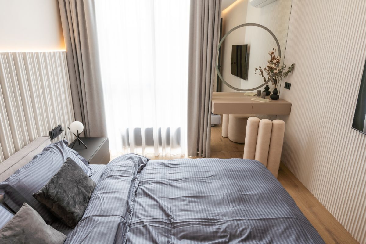 Moderná spálňa so sivou posteľnou bielizňou a minimalistickým toaletným stolíkom pri bielom lamelovom obklade steny.