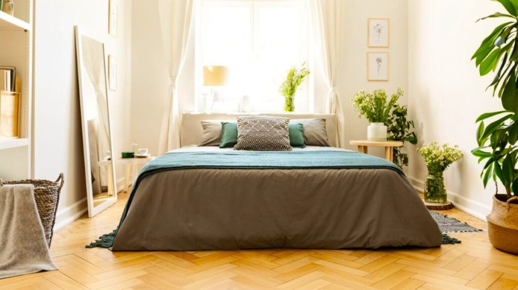 Posteľ v malej spálni so sivou a zelenou bielizňou, opretým zrkadlom na zemi a stolíkom s kvetmi.