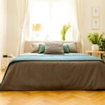 Posteľ v malej spálni so sivou a zelenou bielizňou, opretým zrkadlom na zemi a stolíkom s kvetmi.