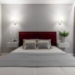 Elegantná posteľ s vínovočerveným zamatovým čelom a bielymi nástennými stolíkmi pred vzorovanou sivou tapetou.