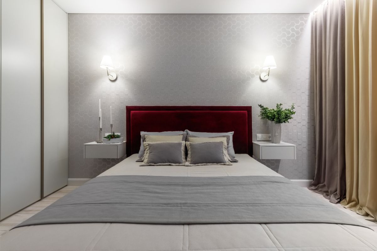 Elegantná posteľ s vínovočerveným zamatovým čelom a bielymi nástennými stolíkmi pred vzorovanou sivou tapetou.