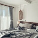 Svetlá spálňa s masívnym dreveným čelom postele a veľkým prúteným lustrom vedľa bielych nariasených záclon.