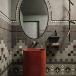 Oválne zrkadlo nad terakotovým betónovým umývadlom v kúpeľni s mozaikovým obkladom.