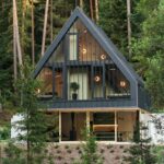 Moderná chata v tvare A s čiernou plechovou fasádou, preskleným prízemím a svetlým dreveným obkladom stien uprostred lesa.