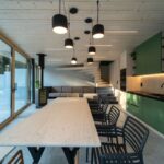 Interiér so svetlým dreveným stropom, čiernymi závesnými lampami, dreveným schodiskom a presklenou stenou s výhľadom na les.